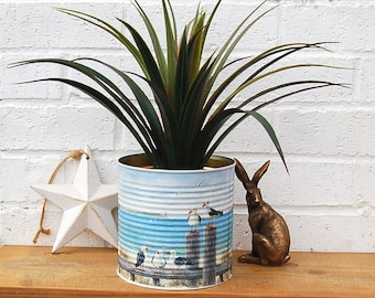 May include: A white metal tin can with a seagull design is filled with a green plant. The can is sitting on a wooden surface with a white star and a brown rabbit figurine.