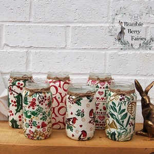 May include: Six glass jars decorated with Christmas-themed fabric, featuring red, green and white patterns. The jars are arranged in a row on a wooden surface, with a gold rabbit figurine to the right.