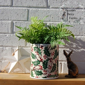 May include: A green and white tin can with a holly and berry pattern is used as a planter for a green fern plant. A white star and a brown rabbit figurine are on a wooden surface in front of the planter.