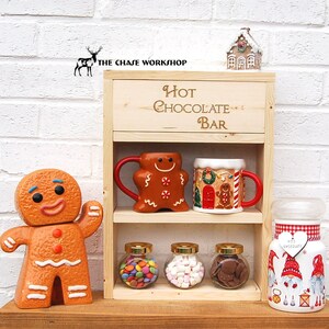 May include: Wooden hot chocolate bar with two shelves. The top shelf has a sign that says "Hot Chocolate Bar". The bottom shelf has three glass jars with candy, marshmallows, and chocolate chips. There are also two mugs with gingerbread designs.