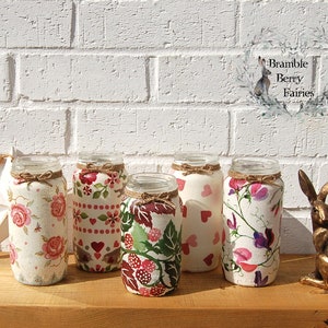 May include: Five glass jars decorated with different floral patterns, including pink roses, red hearts, green leaves, and pink flowers. The jars are arranged on a wooden surface in front of a white brick wall. The text "Bramble Berry Fairies" is visible in the upper right corner of the image.