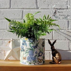 May include: A green fern plant in a white tin can with a floral pattern. The can is sitting on a wooden surface with a white star and a gold rabbit figurine.