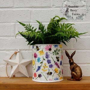 May include: A white metal can with a floral and bee design is used as a planter for a green fern plant. The can is decorated with a watercolour-style pattern of flowers, leaves, and bees. The can is sitting on a wooden surface with a white star and a gold rabbit figurine.