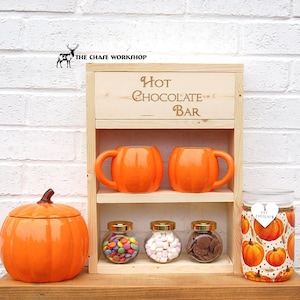 May include: A wooden hot chocolate bar with two orange mugs, three glass jars filled with candy, marshmallows, and chocolate chips, and a pumpkin-themed jar with the text "Hot Chocolate".