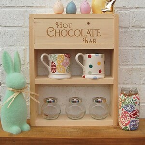 May include: A wooden hot chocolate bar with two shelves. The top shelf has two mugs and a sign that says "Hot Chocolate Bar". The bottom shelf has three glass jars and a jar with a colorful Easter egg design. There are also colorful Easter eggs and a gold bunny on top of the bar.