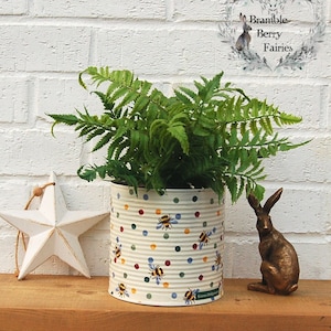 May include: A white ceramic planter with a polka dot pattern and bee illustrations. The planter is filled with a green fern plant. The planter is sitting on a wooden surface with a white star and a bronze rabbit figurine.