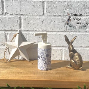 May include: A white star decoration, a soap dispenser with a seashell pattern, and a gold rabbit ornament are arranged on a wooden surface. The soap dispenser features a white pump and a cylindrical body decorated with blue and white seashell designs.