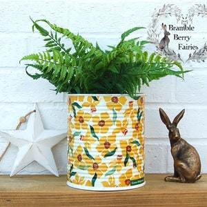 May include: A decorative tin can planter with a yellow floral pattern, filled with green ferns. A white star and a bronze rabbit figurine are on a wooden surface. The background is a white brick wall.
