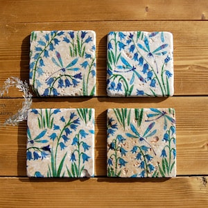 May include: Set of four square coasters featuring a bluebell and dragonfly design. The coasters are made of a light beige stone with a textured surface.