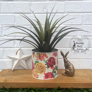 May include: A decorative tin can planter with a floral design, holding a green plant. A gold-coloured rabbit figurine and a white star-shaped ornament are on a wooden shelf against a white brick wall. The planter has a diameter of approximately 15 cm.