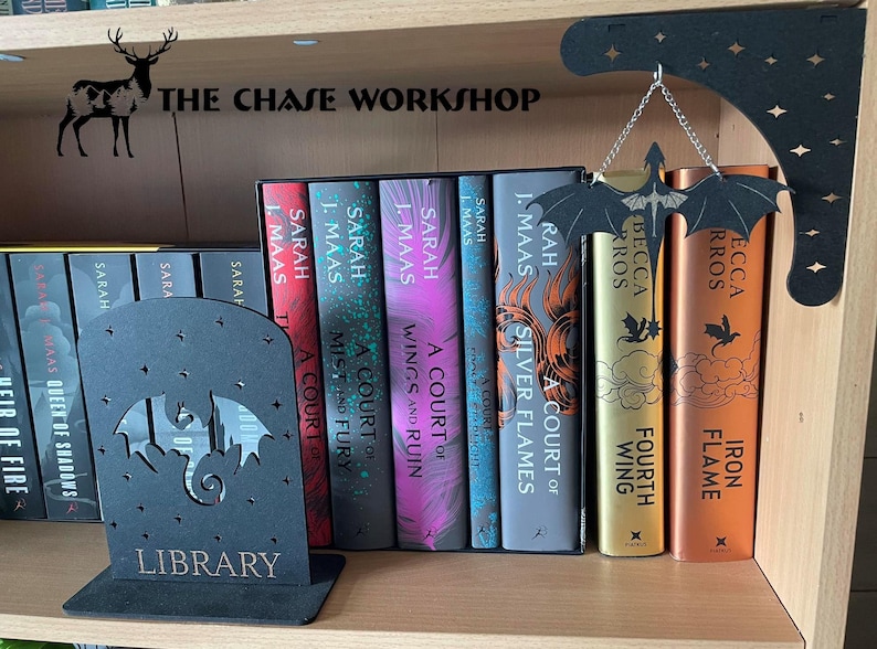 May include: A black metal bookend featuring a dragon silhouette and the inscription "LIBRARY". The bookend is displayed on a bookshelf with a collection of fantasy novels, including "A Court of Silver Flames" and "A Court of Wings and Ruin" by Sarah J. Maas.