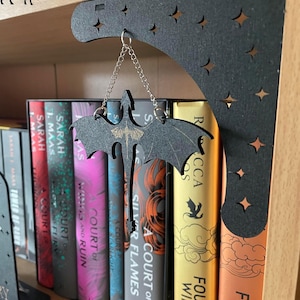 May include: A black metal bookend shaped like a dragon with stars cut out of the design. The bookend is hanging from a chain and is holding up a stack of books with the titles "A Court of Silver Flames", "A Court of Frost and Starlight", "A Court of Wings and Ruin", "A Court of Thorns and Roses", and "A Court of Mist and Fury".