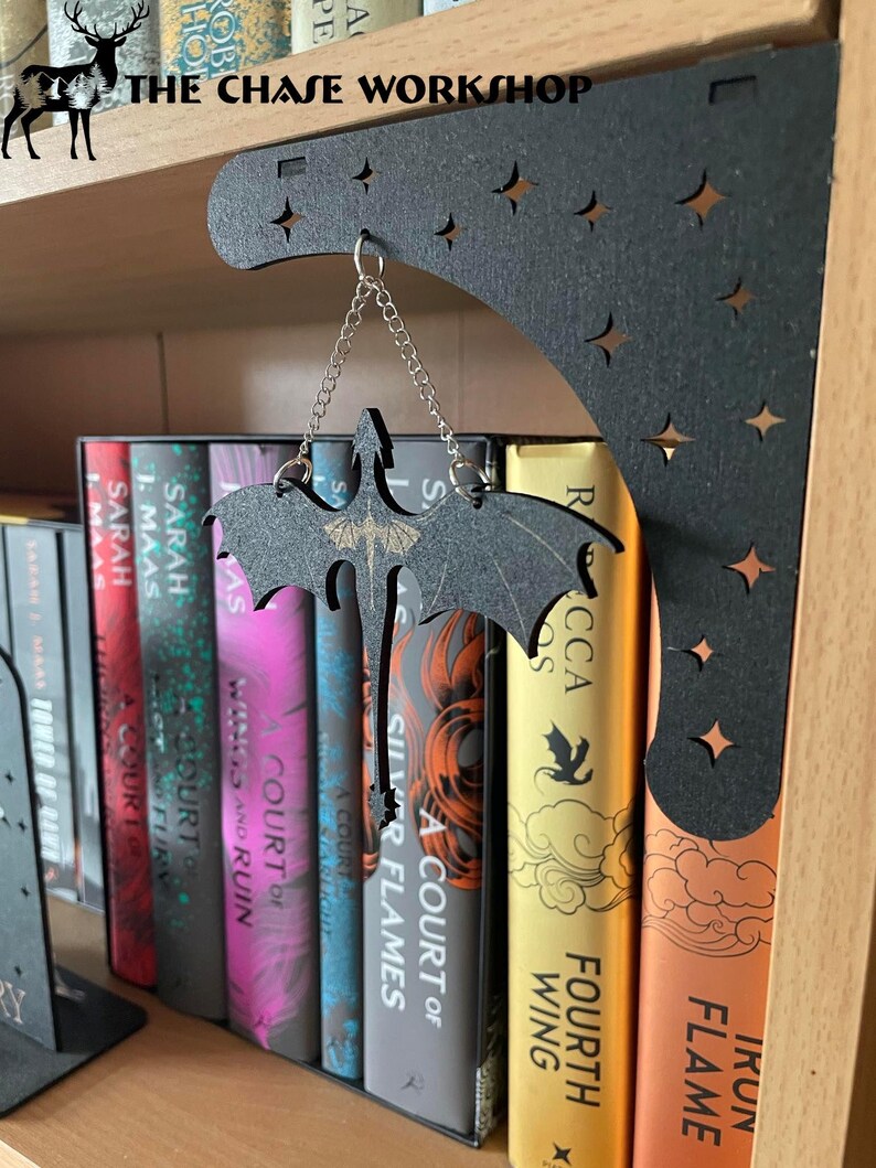 May include: A black metal bookend shaped like a dragon with stars cut out of the design. The bookend is hanging from a chain and is holding up a stack of books with the titles "A Court of Silver Flames", "A Court of Frost and Starlight", "A Court of Wings and Ruin", "A Court of Thorns and Roses", and "A Court of Mist and Fury".