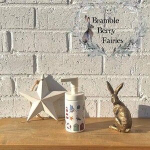 May include: White ceramic soap dispenser with a nautical theme. The dispenser has a pump top and is decorated with images of lighthouses, sailboats, seagulls, and beach houses. The dispenser is sitting on a wooden shelf with a white star and a gold rabbit figurine.