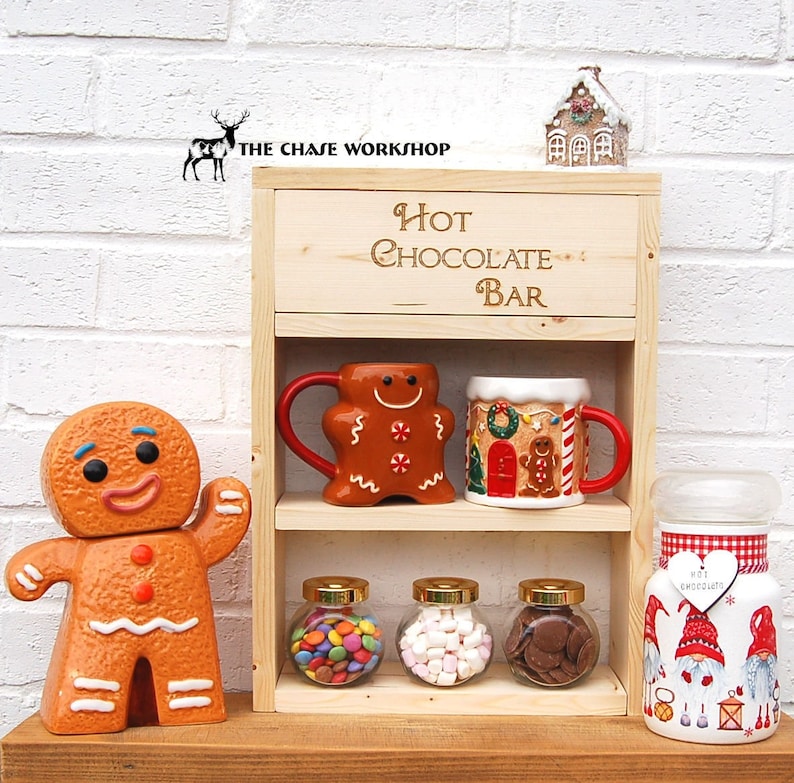 May include: Wooden hot chocolate bar with two shelves. The top shelf has a sign that says "Hot Chocolate Bar". The bottom shelf has three glass jars with candy, marshmallows, and chocolate chips. There are also two mugs with gingerbread designs.