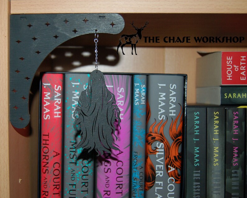 May include: A black wooden shelf bracket with a decorative star pattern. A black silhouette of a hooded figure hangs from the bracket. The bracket is mounted on a wooden shelf with a row of books titled "A Court of Thorns and Roses" by Sarah J. Maas.