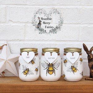 May include: Three white mason jars with gold lids and bee illustrations. Each jar has a white heart-shaped tag with the words "tea", "coffee", and "sugar" written on them. The jars are sitting on a wooden shelf with a white star and a gold rabbit figurine.
