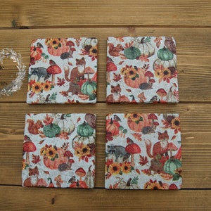 May include: Set of four square coasters featuring a autumnal design. The coasters have a white background with a pattern of pumpkins, sunflowers, mushrooms, leaves, and woodland creatures. The coasters are made of cork and have a rustic appearance.