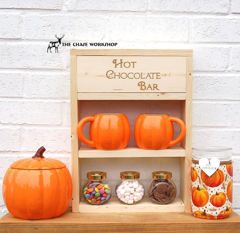 May include: A wooden hot chocolate bar with two orange mugs, three glass jars filled with candy, marshmallows, and chocolate chips, and a pumpkin-themed jar with the text "Hot Chocolate".