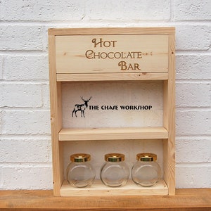 May include: Wooden shelf with the words "Hot Chocolate Bar" and "The Chase Workshop" engraved on it. The shelf has three glass jars with gold lids.
