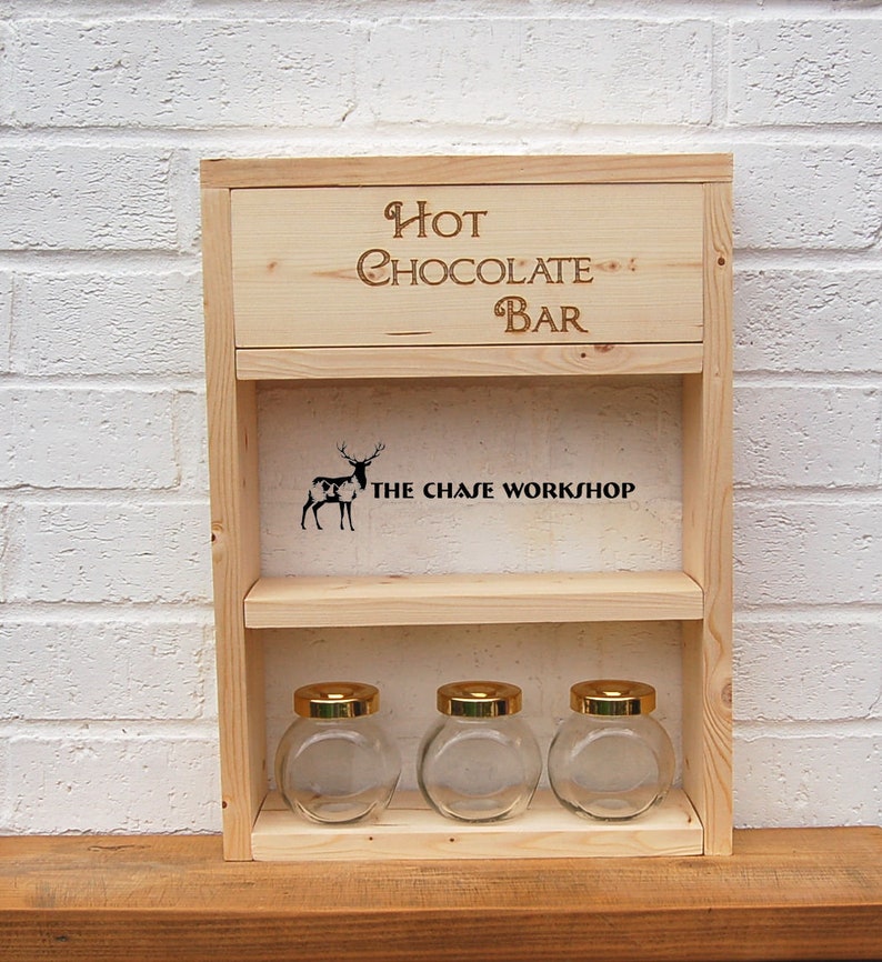 May include: Wooden shelf with the words "Hot Chocolate Bar" and "The Chase Workshop" engraved on it. The shelf has three glass jars with gold lids.