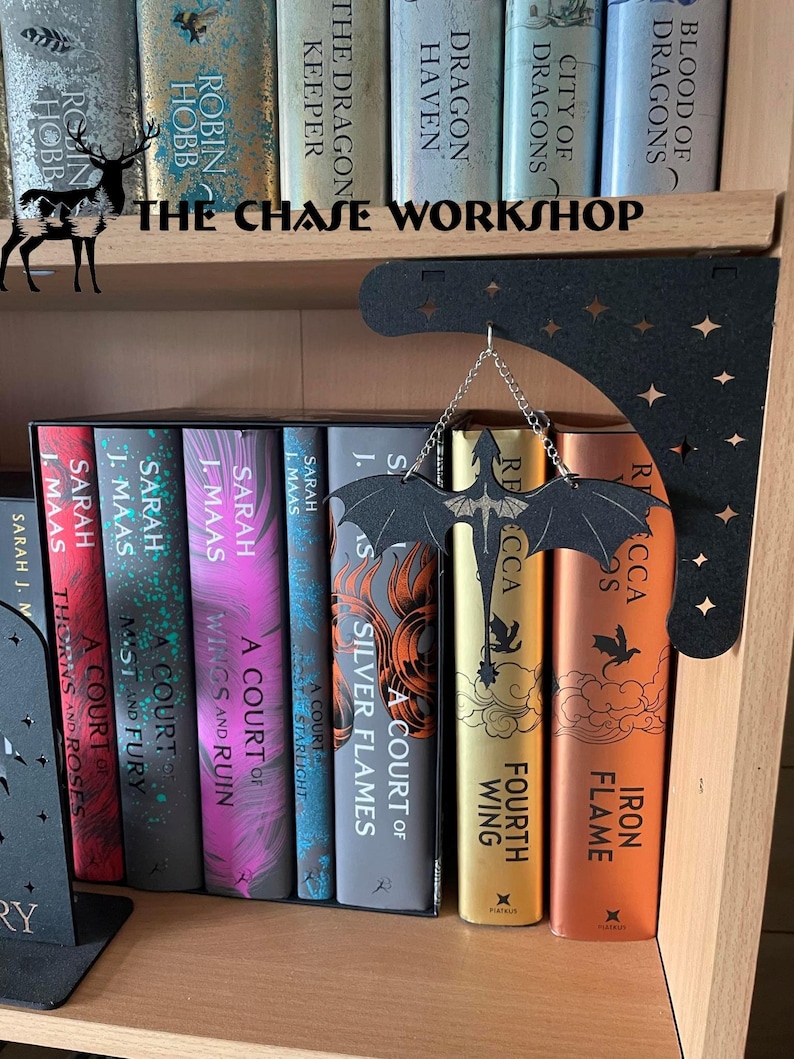 May include: A bookshelf with a black metal dragon bookend and a black metal crescent moon hanging from the top shelf. The bookshelf is filled with fantasy novels, including the "A Court of Thorns and Roses" series by Sarah J. Maas and the "Dragon" series by Margaret Weis and Tracy Hickman.