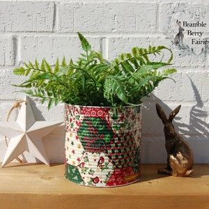 May include: A green fern plant in a tin can planter with a patchwork design featuring red, green, white, and gold colours. The planter is decorated with a floral pattern and small birds. The plant is sitting on a wooden surface with a white star and a gold rabbit figurine.