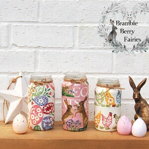 May include: Three glass jars decorated with Easter-themed patterns. The jars are filled with pink, white, and yellow floral designs, including eggs, chickens, and bunnies. The jars are arranged on a wooden surface with two pink speckled eggs and a gold bunny figurine.