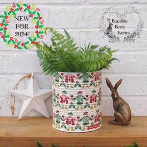 May include: A white metal planter with a Christmas-themed design featuring red and green illustrations of people wearing sweaters and holding hands. The planter is filled with green ferns and is sitting on a wooden surface. A white star ornament and a bronze rabbit figurine are also visible in the image.