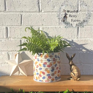 May include: A white metal planter with a colourful polka dot pattern, filled with a green fern plant. The planter is sitting on a wooden surface with a gold rabbit figurine and a white star-shaped decoration.