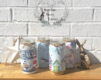 May include: Four decorative glass jars with beach-themed designs, including a lighthouse, sailboat, and beach huts. Each jar is wrapped with patterned paper and tied with twine. A gold-colored rabbit statue and a white star are also present.