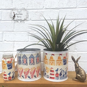 May include: Three decorative tin cans and a jar with beach-themed designs. The cans feature illustrations of beach huts, sandcastles, and nautical elements in red, blue, and yellow. A bronze rabbit figurine sits on a wooden shelf.