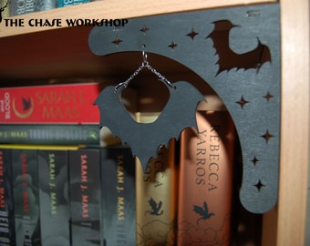 May include: A black wooden bat-shaped shelf bracket with a crescent moon design and stars. A silver chain with a bat-shaped pendant hangs from the bracket. The bracket is mounted on a wooden shelf with books behind it.