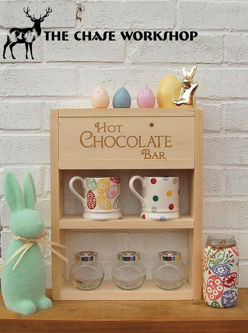 May include: A wooden hot chocolate bar with two shelves. The top shelf has two mugs and a sign that says "Hot Chocolate Bar". The bottom shelf has three glass jars and a jar with a colorful Easter egg design. There are also colorful Easter eggs and a gold bunny on top of the bar.
