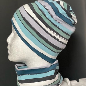 Pode incluir: Um gorro azul, branco e cinza listrado com um cachecol combinando. O gorro é feito de um tecido macio e elástico e tem um caimento folgado.