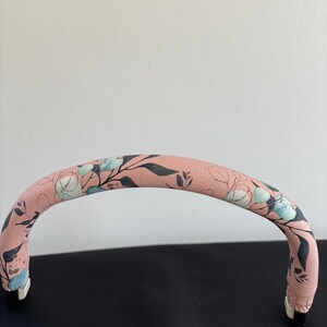 Könnte beinhalten: Ein rosa Kinderwagen-Griffbezug mit Blumenmuster. Der Bezug hat einen hellrosa Hintergrund mit blauen und weißen Blumen und Blättern.