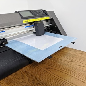 May include: A gray vinyl cutter with a blue cutting mat and a white sheet of paper. The machine has a control panel with buttons and a small screen. The cutting mat is on a wooden surface.