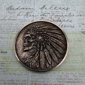May include: A bronze-colored coin featuring a detailed relief of a skull wearing a Native American headdress. The coin has a circular shape with intricate feather and star details, set against a textured background.