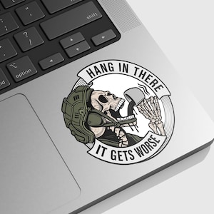 May include: A sticker featuring a skeleton wearing a helmet and drinking from a mug. The sticker has the text "HANG IN THERE" above the skeleton and "IT GETS WORSE" below. The sticker is on a laptop.