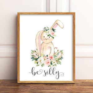 May include: A watercolor illustration of a beige bunny wearing a pink and white flower crown. The bunny is sitting on a bed of pink and white flowers with green leaves. The text "be silly" is written below the bunny.
