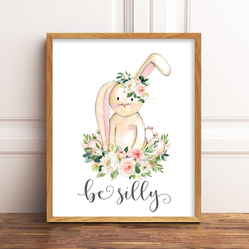 May include: A watercolor illustration of a beige bunny wearing a pink and white flower crown. The bunny is sitting on a bed of pink and white flowers with green leaves. The text "be silly" is written below the bunny.