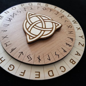 Peut inclure: Une roue de chiffrement en bois avec un motif de nœud celtique au centre. La roue comporte trois couches, avec des lettres et des runes gravées. La couche supérieure présente une finition marron clair, tandis que les couches inférieures sont d'une teinte plus claire.