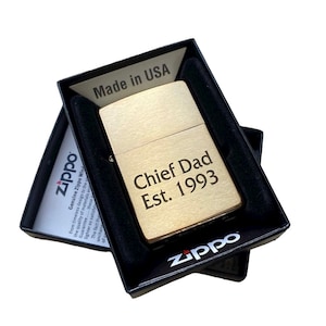 Puede incluir: Un encendedor Zippo dorado con la inscripci&oacute;n "Chief Dad Est. 1993" en una caja negra.