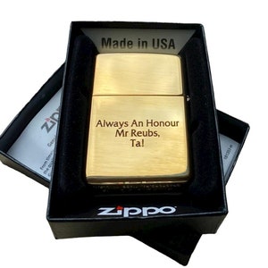Puede incluir: Un encendedor Zippo dorado con acabado cepillado y un mensaje grabado que dice "Always An Honour Mr Reubs, Ta!" El encendedor est&aacute; en una caja negra con el logotipo de Zippo.