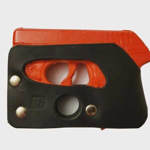 Puede incluir: Una pistola naranja est&aacute; parcialmente encerrada en una funda de cuero negro. La funda est&aacute; asegurada con tres remaches plateados y presenta una abertura circular y un logotipo "S" estilizado. El gatillo y la empu&ntilde;adura de la pistola son visibles.