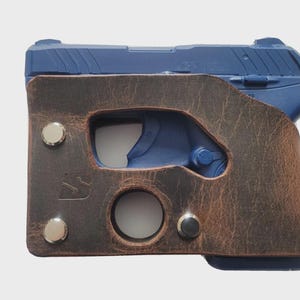 Puede incluir: Una pistola azul est&aacute; parcialmente encerrada en una funda de cuero marr&oacute;n. La funda tiene un acabado envejecido, con remaches plateados y una abertura circular. La empu&ntilde;adura de la pistola es visible y la funda tiene un recorte para el gatillo.