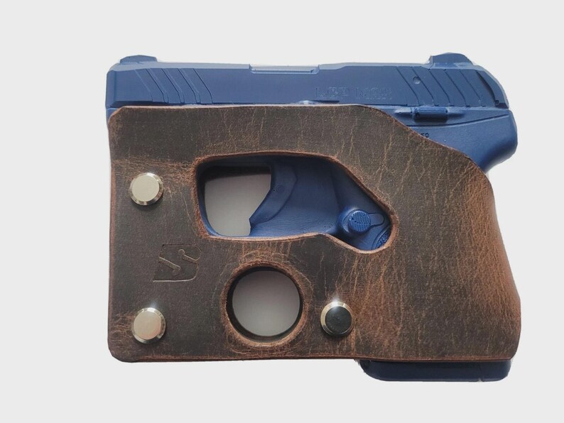 Puede incluir: Una pistola azul est&aacute; parcialmente encerrada en una funda de cuero marr&oacute;n. La funda tiene un acabado envejecido, con remaches plateados y una abertura circular. La empu&ntilde;adura de la pistola es visible y la funda tiene un recorte para el gatillo.
