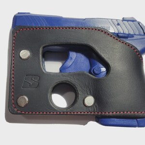Puede incluir: Una funda de cuero negro con costuras rojas y remaches plateados, dise&ntilde;ada para una pistola azul. La funda presenta un recorte para el gatillo y una abertura circular.