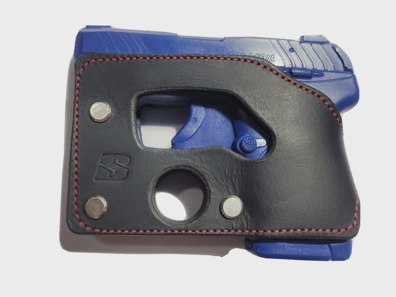 Puede incluir: Una funda de cuero negro con costuras rojas y remaches plateados, dise&ntilde;ada para una pistola azul. La funda presenta un recorte para el gatillo y una abertura circular.