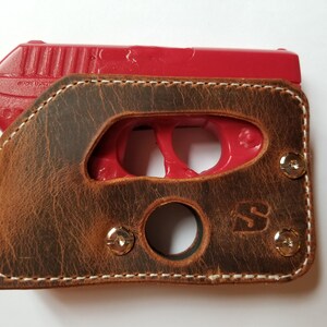 Puede incluir: Una funda de cuero marr&oacute;n con costuras blancas, que sostiene una pistola de entrenamiento roja. La funda tiene un acabado envejecido, remaches met&aacute;licos y un recorte para el gatillo. El cuero est&aacute; estampado con un logotipo.
